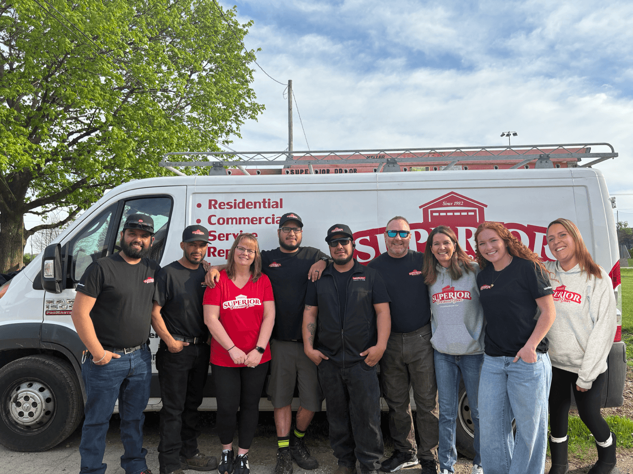 Nine people pose together and smile in front of a white service van with red Superior branding and a ladder on top. The group appears to be work colleagues, wearing branded shirts and standing outdoors on a sunny day.